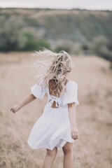 A girl with blond curly hair and in a romantic dress stands in the middle of the field. The wind...