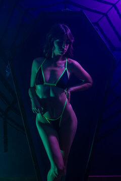 fit thin brunette female wearing a black and lime green bikini in front of a purple and black toe pintcher cofin with smoke colored by green and purple lights