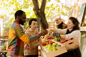 Young diverse clients tasting free apple samples at farmers market, seasonal juicy fruits and vegetables outside. Man and woman purchasing healthy products from local farm marketplace.