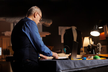 Experienced skilled old tailor following plans to honor customer comission in professional atelier shop workspace. Meticulous focused suitmaker crafting menswear bespoken collection