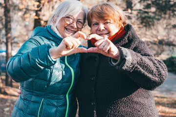 A smiling couple of elderly women hug each other in the park forming a heart with their hands...