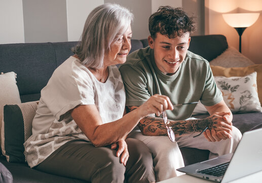 Senior woman and young grandson watch laptop together at home. Young boy teaches computer technology to his elderly grandmother how to surf, buy on internet. Elderly woman learns how to use computer.
