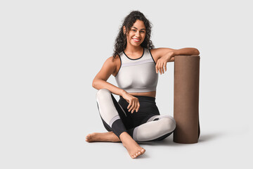 Sporty young African-American woman with yoga mat sitting on grey background