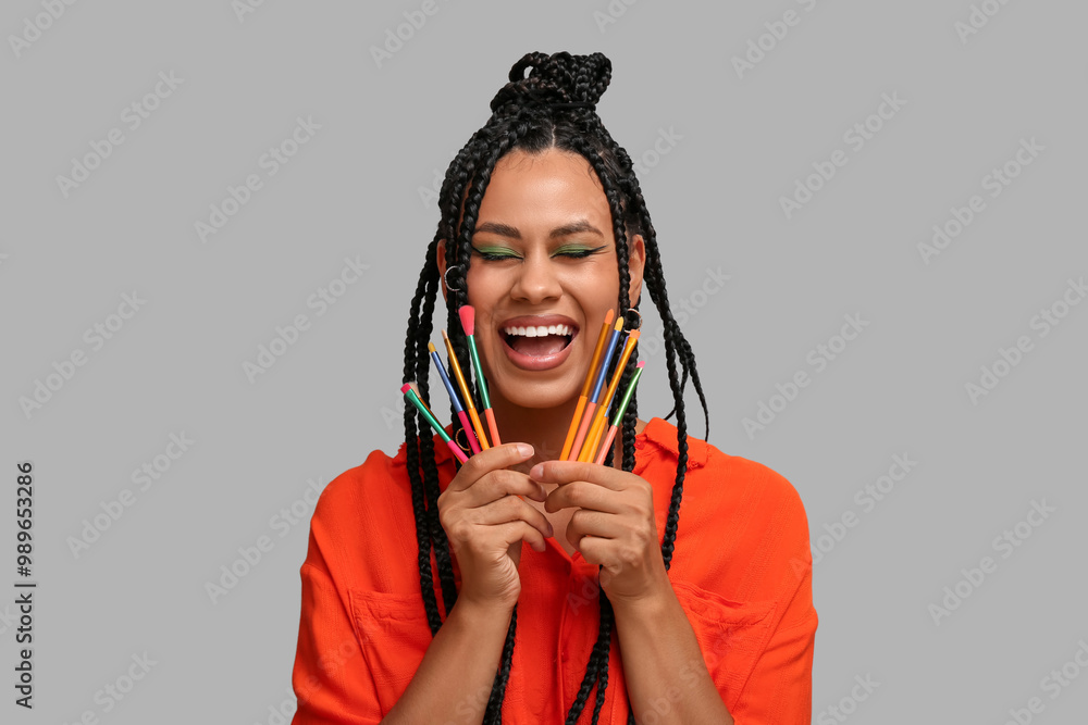 Poster Happy young African-American woman with braids and beautiful makeup holding brushes on grey background