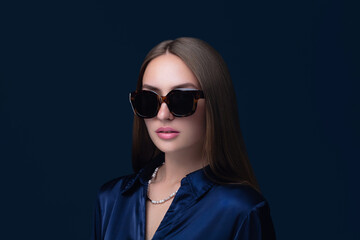 Elegant woman wearing oversized tortoiseshell sunglasses, navy satin blouse, and a pearl necklace. She have a classy, modern style with a minimalistic dark background,  chic fashion look.