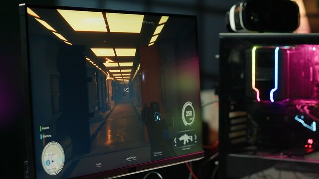 Computer screen showing first person shooter game left open at night in empty apartment with neon lights. Close up of PC on desk in living room with no people in and display left open