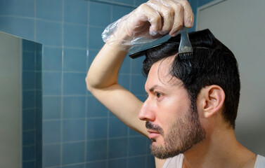 Hombre tiñéndose el pelo y patilla en baño de casa, usando cepillo o brocha, guante y peine.  