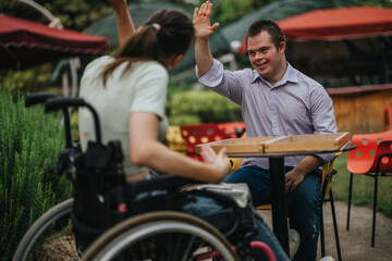 A boy with Down syndrome high-fives a girl in a wheelchair while playing backgammon at an outdoor table. The scene captures a moment of joy and inclusivity.