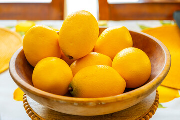 A stack of ripe lemons in a wooden bowl on a dining table with sunlight coming through windows behind.