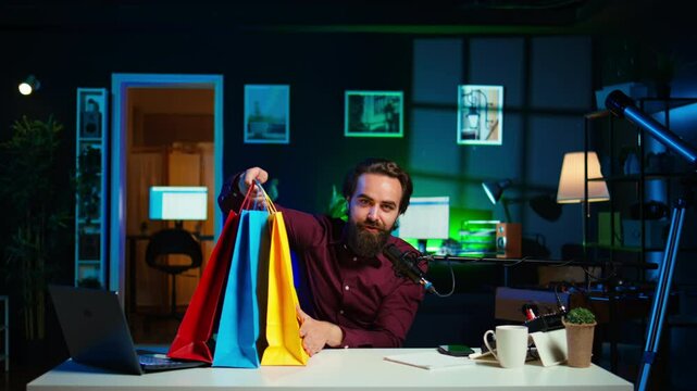 Upbeat influencer holding colorful shopping bags in hand, showing subscribers purchases he recently got. Cheerful content creator presenting fanbase new acquisitions, camera B