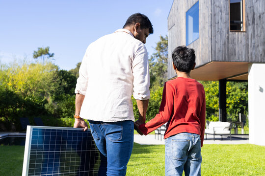 indian father and son carrying solar panel, walking together in backyard