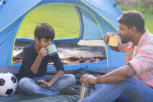 Camping outdoors, Indian father and son drinking from mugs near tent and soccer ball