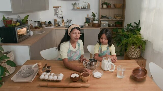 High angle shot of Latin aunt in hairband and apron sitting at table with small niece and showing ingredients for homemade pastry, explaining process of making dough at home kitchen