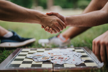 Obraz premium Two people shaking hands over a chessboard with scattered playing cards, symbolizing agreement, strategy, and teamwork in an outdoor environment.