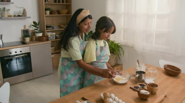 Side shot of 10-year-old Hispanic girl in apron learning how to break eggs while making dough for homemade pastry together with kind aunt at home