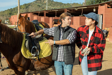 Two positive male female grooms talking and checking saddle on brown horse for riding in yard of...