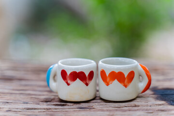 Two small, white ceramic mugs with colored heart designs sit side-by-side on a weathered wooden table. The background is blurred, with a soft green color suggesting a natural outdoor setting.