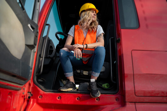 Young woman truck driver with safety vest and hard hat sitting on the drivers seat with doors open, resting 