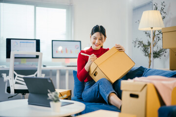 Young asian woman is sitting on her couch and writing on a cardboard box, preparing it for shipping. She is working from home