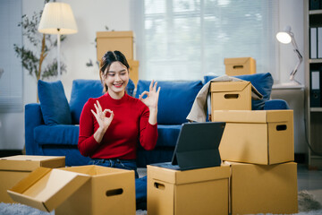 Young woman is having a video call on her tablet and making the ok sign with both hands while sitting on the floor of her new apartment, surrounded by cardboard boxes