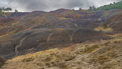 The unique 23 coloured earth of Mauritius. Colorful sand hills against a cloudy sky. Green vegetation is nearby. La Vallée Des Couleurs Nature Park