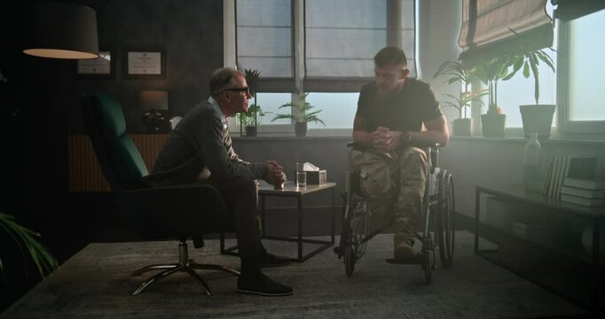 Senior psychotherapist conducts psychological therapy or post war rehabilitation for military man in wheelchair. Psychiatrist talks, gives sedative drugs or antidepressants to soldier with disability.