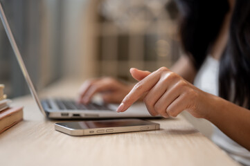 A female's finger tapping on a phone screen on a table while working on her laptop in a coffee shop