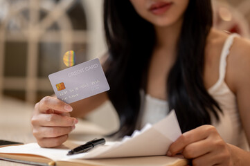 A thoughtful Asian woman holding a credit card and reviewing receipts at a table indoors.