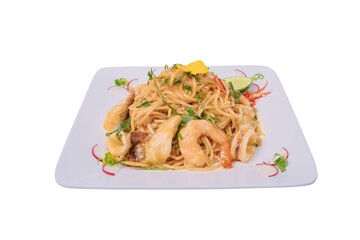 A square plate holds a flavorful stir-fried spaghetti dish with seafood, vegetables, and a spicy sauce, garnished with a flower and lime wedge.