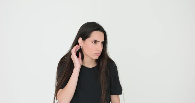 Curious woman with long loose dark hair holds hand next to ear trying to hear gossiping on white background. Female looks checking earring