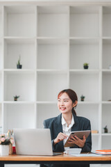 A young professional woman multitasking with a laptop and tablet in a modern office setting, featuring a background of shelves with plants and decor.