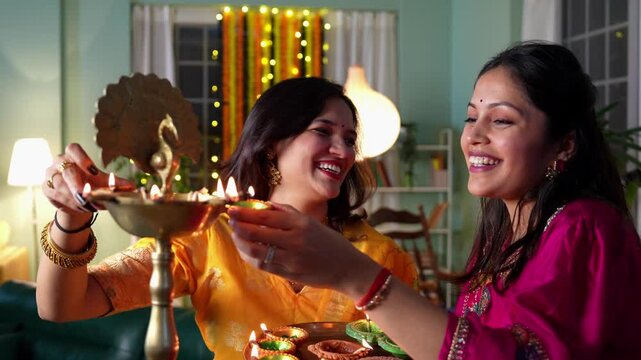 Two beautiful Indian women in traditional bright clothes and jewellery light Samai with oil diyas welcoming Diwali with smiles at home adorned with stunning festive decorations