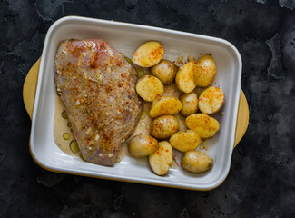 Cooking lunch - raw marinated chicken breast and baby potatoes in a baking dish on a dark background, top view