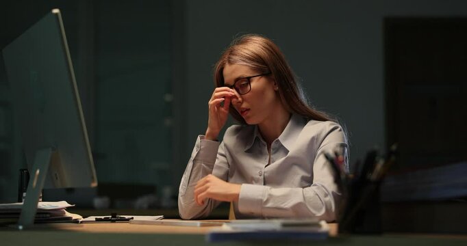 Exhausted businesswoman massages temples seeking relief from headache at computer. Late night work creates stress and pressure at workplace