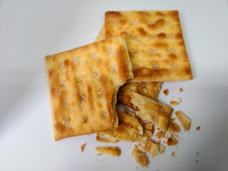 A few crispy crackers, one partially eaten, lie on a white surface with visible crumbs. The image showcases the flaky texture, golden color, and sugar crystals, emphasizing a snack in mid-bite.