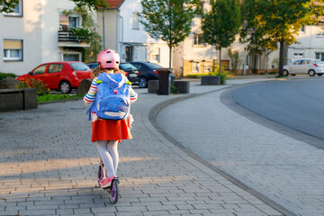 Little happy girl on way to school by pushing scooter. Elementary school child riding in the city, with big satchel. Kid with pink helmet. Safe route to school and movement for children concept.