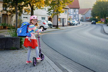 Little happy girl on way to school by pushing scooter. Elementary school child riding in the city, with big satchel. Kid with pink helmet. Safe route to school and movement for children concept.