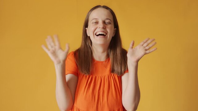 Cheerful woman with brown hair wearing orange shirt greeting with wave and big smile expressing happiness and friendliness welcomes everyone isolated over yellow background