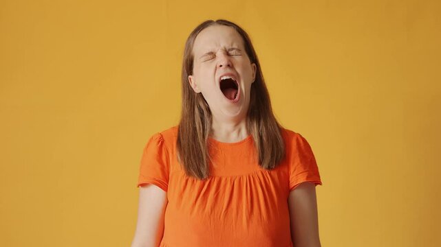 Sleepy woman with brown hair wearing orange shirt yawning and showing tired expression dealing with exhaustion and boredom isolated over yellow background