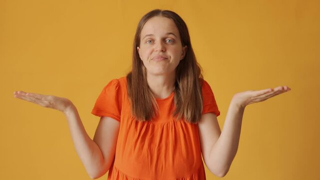 Confused woman with brown hair wearing orange shirt looking unsure and ambiguous raising her arm in contemplation about decision and displaying copy space isolated over yellow background