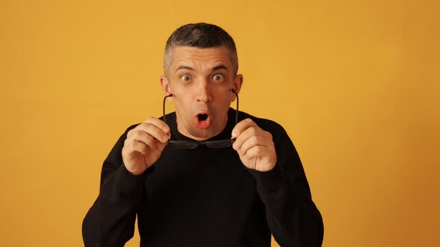 Focused man wearing glasses and black raglan checking eyesight while taking off eyeglasses after vision correction sees everything without spectacles isolated over yellow background