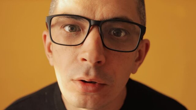 Closeup of surprised man in glasses wearing black jumper displaying shocked expression posing with open mouth hearing unexpected news or triumph isolated over yellow background