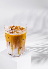 Iced caramel latte standing on white cube with palm leaf shadow
