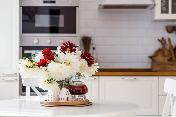 Still-life. White and red dahlia flowers in a vase, a teapot and white houses on a white table in the interior of a home kitchen. A cozy autumn concept.
