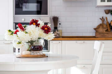 Still-life. White and red dahlia flowers in a vase, a teapot, a pumpkin and a house on a white table in the interior of a home kitchen. A cozy autumn concept.