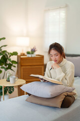 Young asian women in sweater sitting on comfortable couch and reading a book for thinking and...