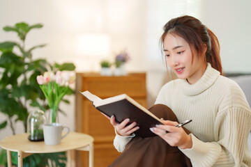 Young asian women in sweater reading the book to learning about knowledge and education while sitting on couch to relaxation and doing activity for spending time with slow life lifestyle at home