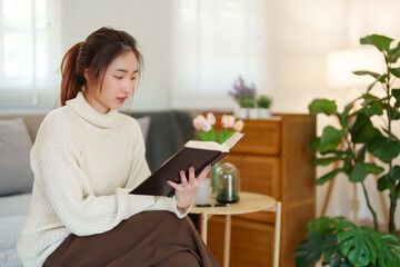 Young asian women in sweater reading a book and learning about literature story with enjoying while sitting on couch to relaxation and doing activity to spending time with slow life lifestyle at home