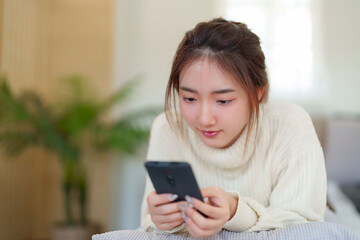 Young asian women in sweater lying on comfortable couch to enjoying with chatting social media on smartphone while relaxation and doing activity to spending time with slow life lifestyle at home