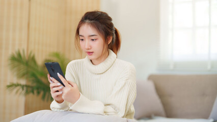 Young asian women in sweater enjoying with surfing entertainment on smartphone while sitting on comfortable couch to relaxation and doing activity for spending time with slow life lifestyle at home